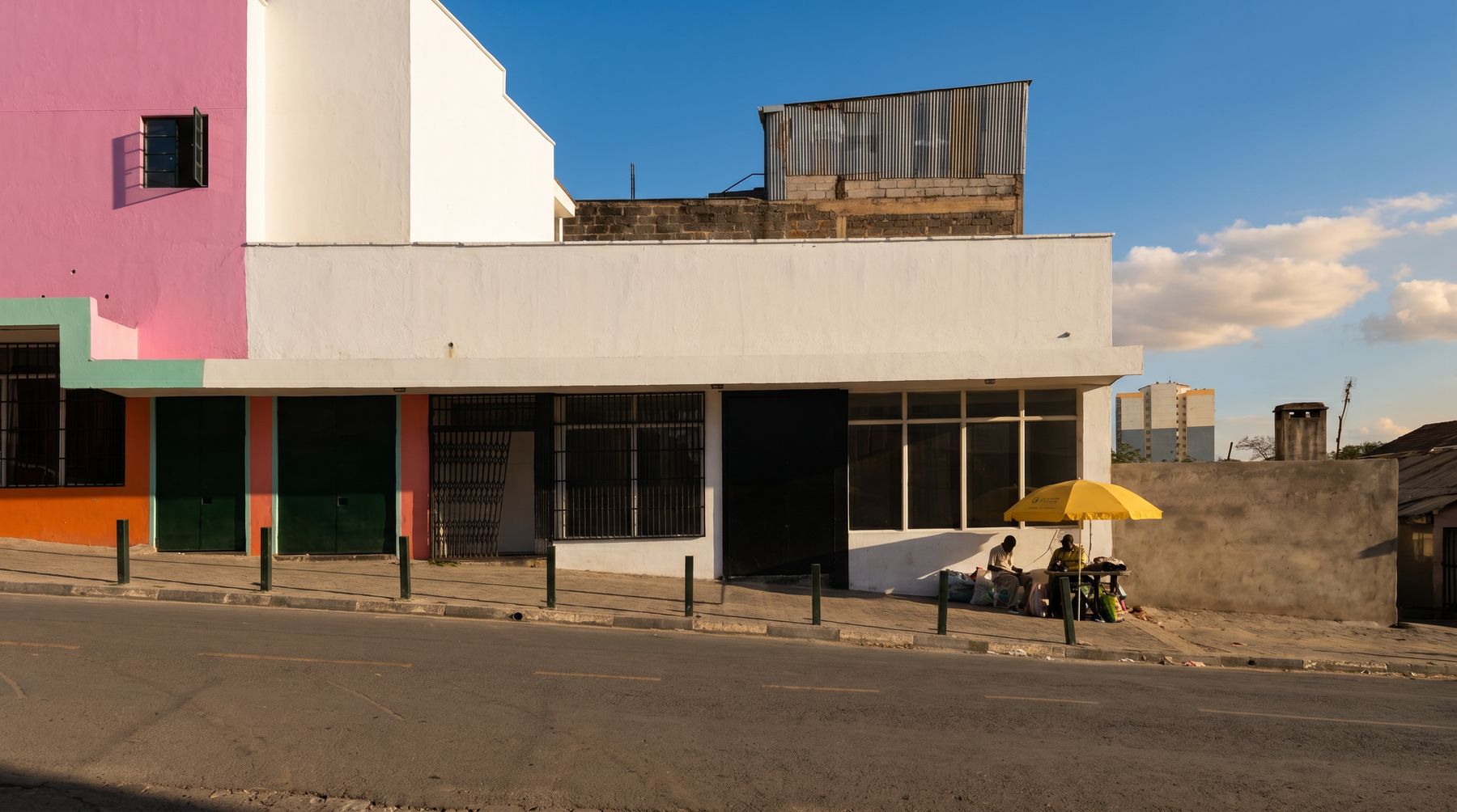 The Ngara Food Projects building on its street in Ngara, Nairobi: a pink and white facade with green doors, and neighbours at a stall under a yellow umbrella.