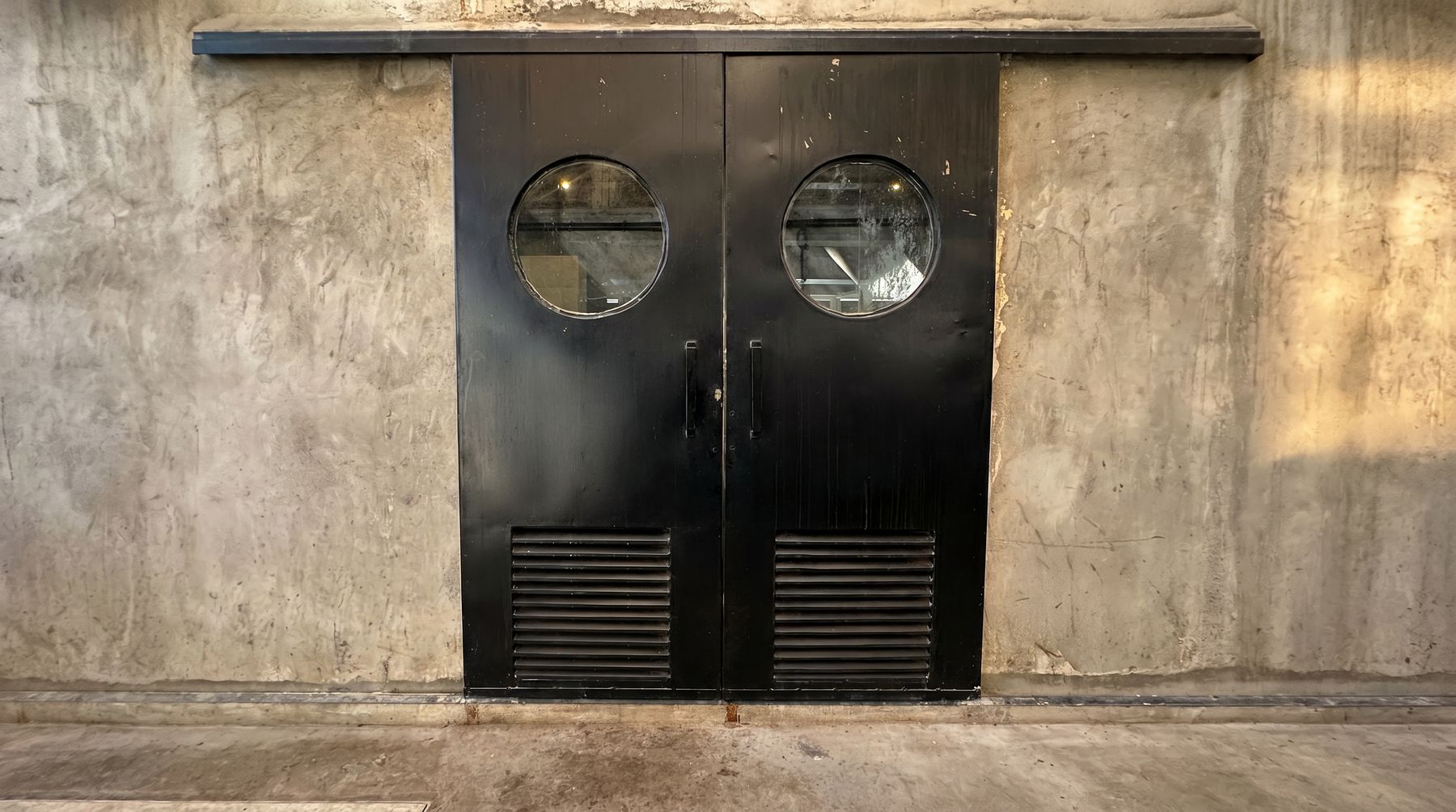 The kitchen's black double doors with round porthole windows, set in a raw concrete wall.